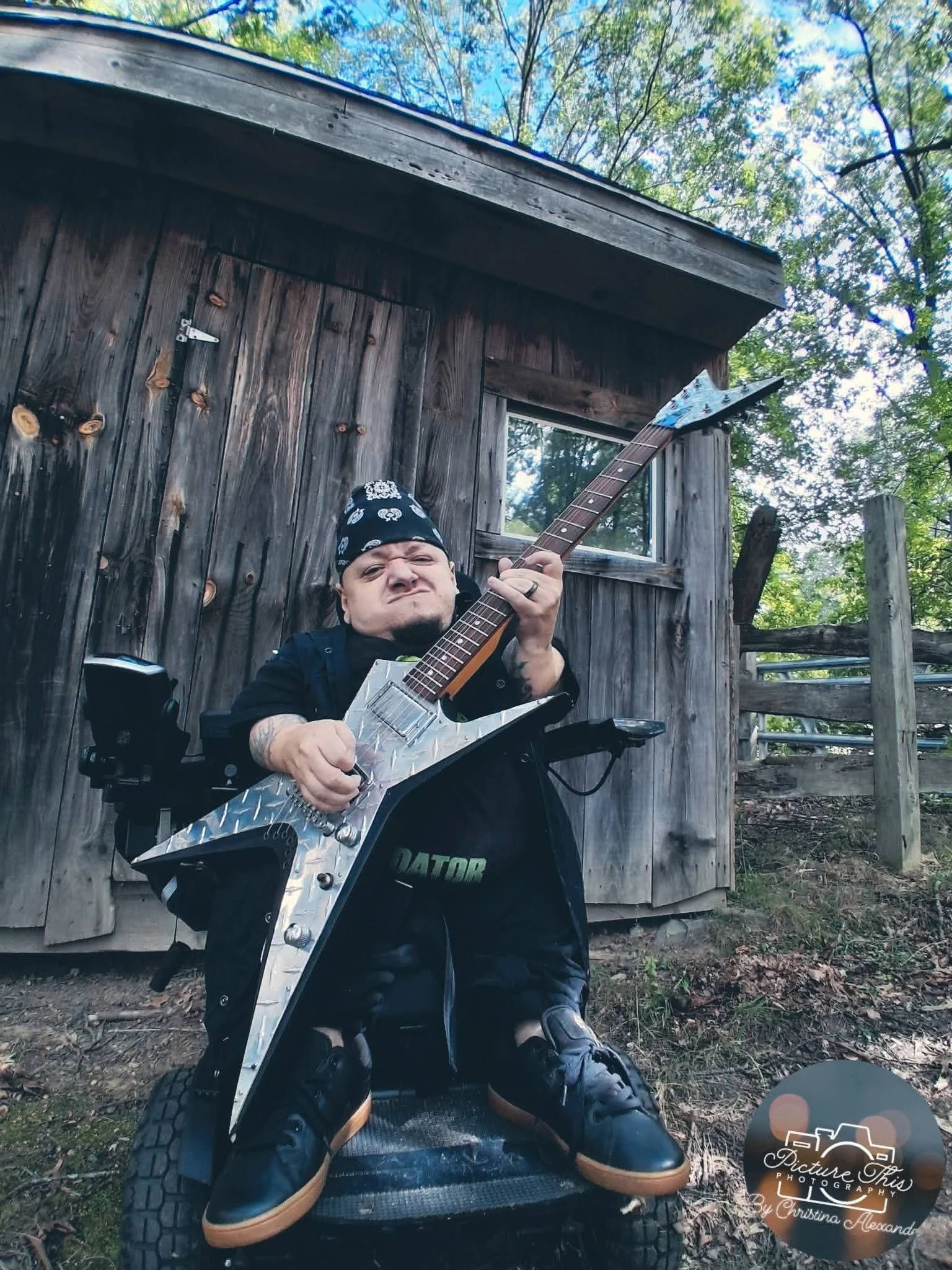 Josh playing a silver electric guitar in front of a rustic wooden cabin. The photographer watermark remains visible in the lower-right corner.